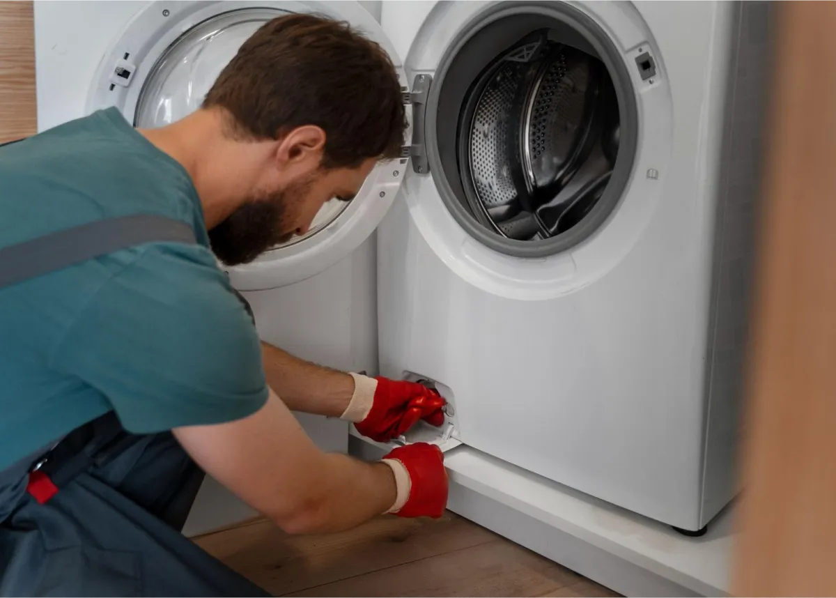 Appliance repair technician in teal shirt and red work gloves crouching to service the drain connection